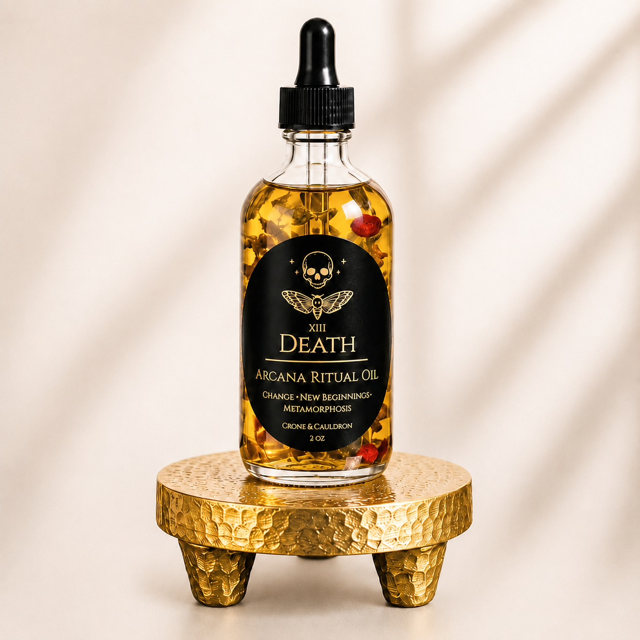 A clear glass dropper bottle filled with golden ritual oil and containing dried botanicals, resting on a small hammered gold pedestal. The bottle has a black label with a skull logo and the brand name 'XIII DEATH'.