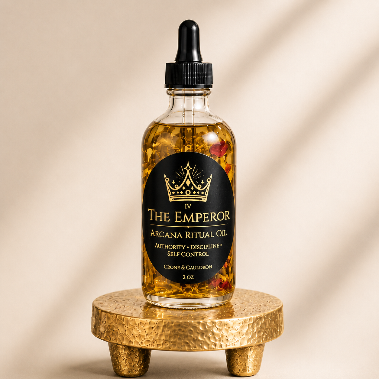 A 2 oz glass bottle of 'The Emperor' Arcana Ritual Oil, filled with golden oil and dried botanicals, resting on a small hammered gold stand against a warm beige background with soft diagonal shadows.