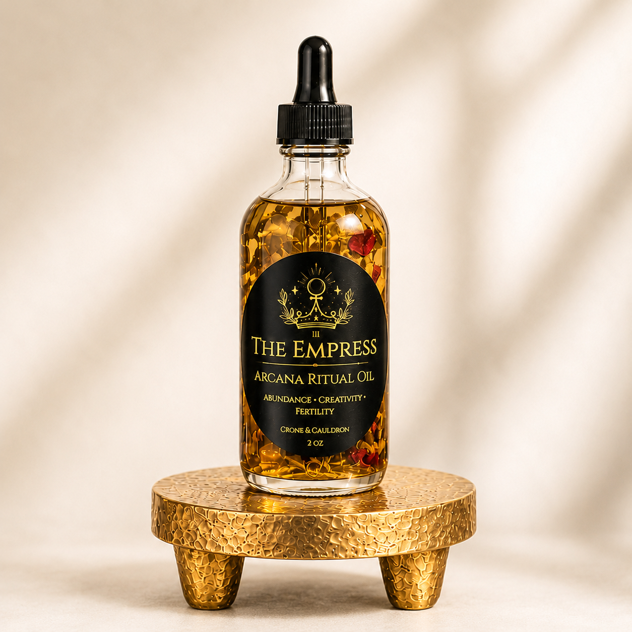 A 2 oz glass bottle of The Empress Arcana Ritual Oil with a black dropper cap, filled with golden oil and dried botanicals, displayed on a small textured gold stand against a warm beige background.
