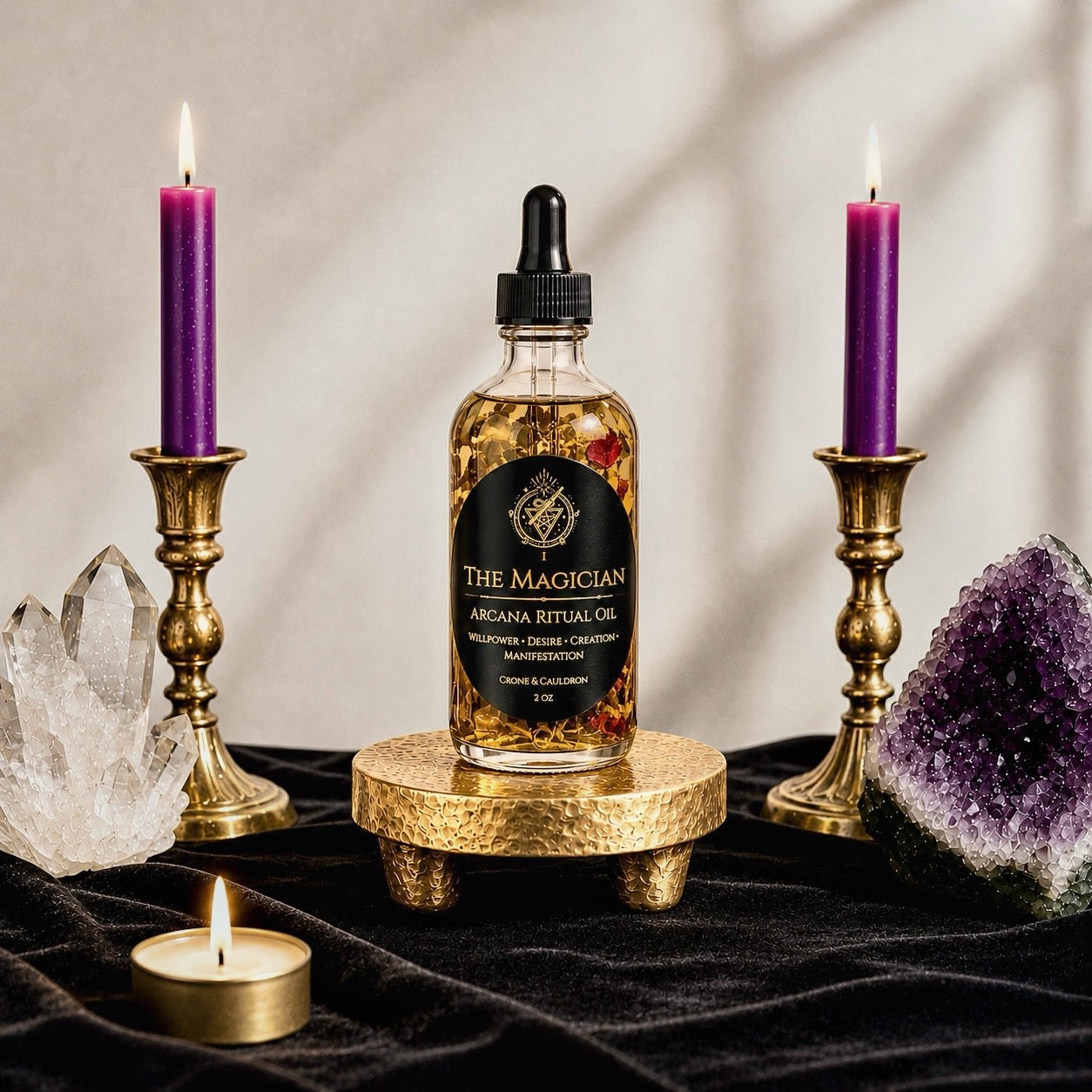 A bottle of The Magician Arcana Ritual Oil sits on a small gold pedestal, flanked by two lit purple candles in gold holders, with crystal clusters and a small tea light on a black velvet surface.