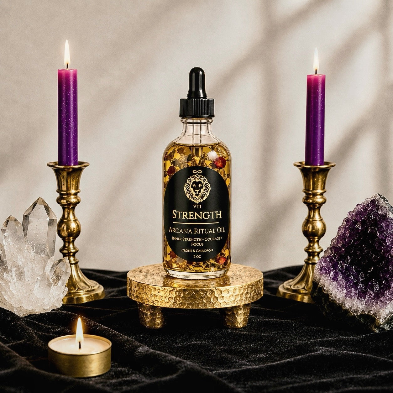 A bottle of 'The Strength' Arcana Ritual Oil sits on a small gold pedestal, flanked by two lit purple candles in gold holders, with crystals and a small tea light on a black velvet surface.