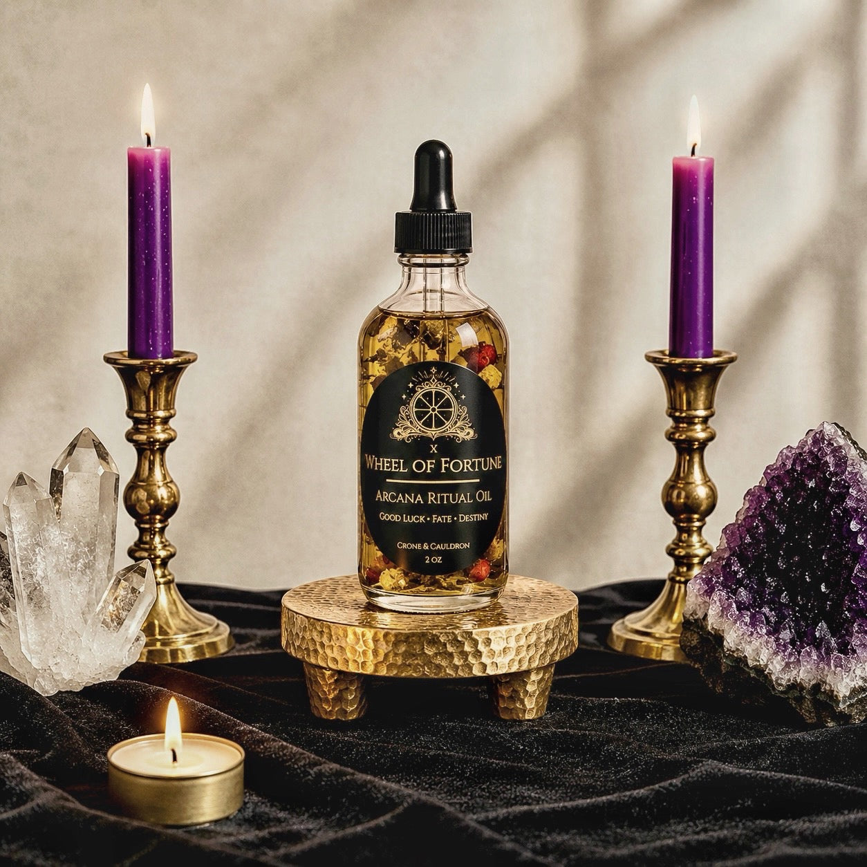 A bottle of Wheel of Fortune Arcana Ritual Oil sits on a small gold pedestal, flanked by two lit purple candles in gold holders, with crystals and a small tea light on a dark draped surface.