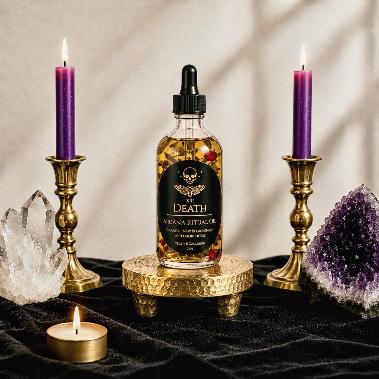 A bottle of Death Arcana Ritual Oil stands on a small gold pedestal, flanked by two lit purple candles in gold holders, with crystals and a small tea light on a black cloth against a softly lit beige background.