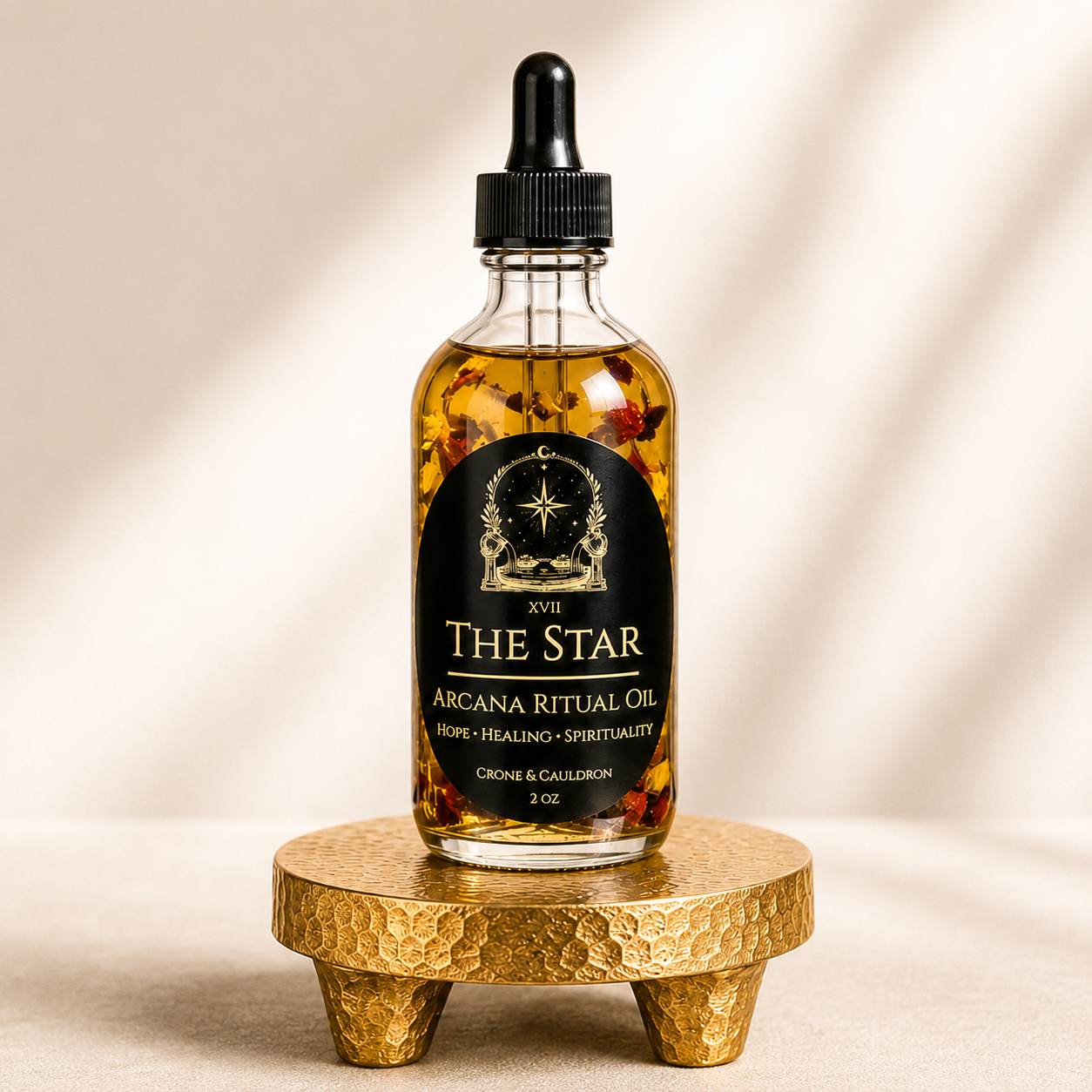 A 3 oz glass dropper bottle of XVII The Star Arcana Ritual Oil, filled with golden oil and dried botanicals, displayed on a small hammered gold stand against a warm beige background with soft shadows.