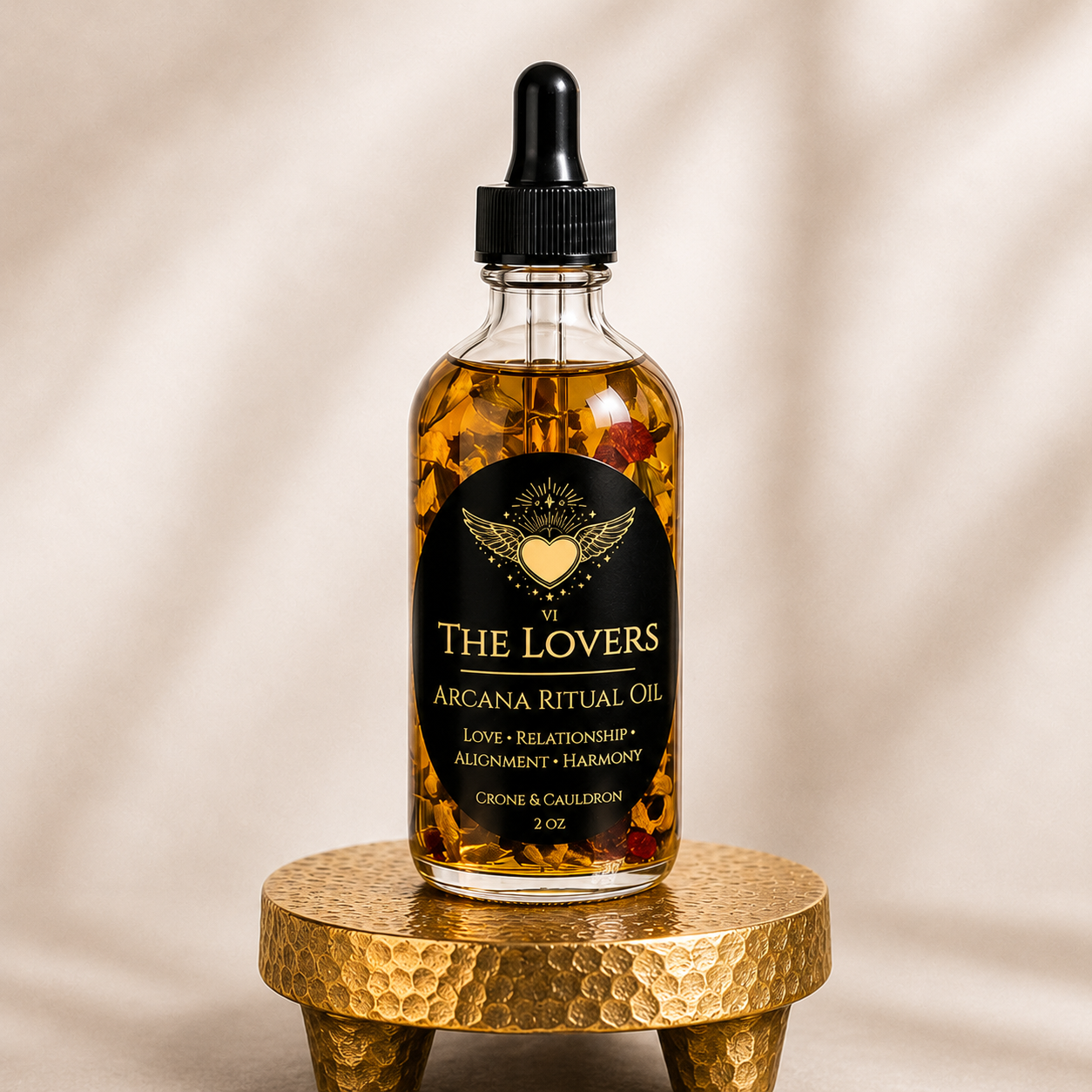 A 2 oz glass bottle of 'The Lovers' Arcana Ritual Oil with a black dropper cap, resting on a textured gold pedestal against a warm beige background with soft leaf shadows.